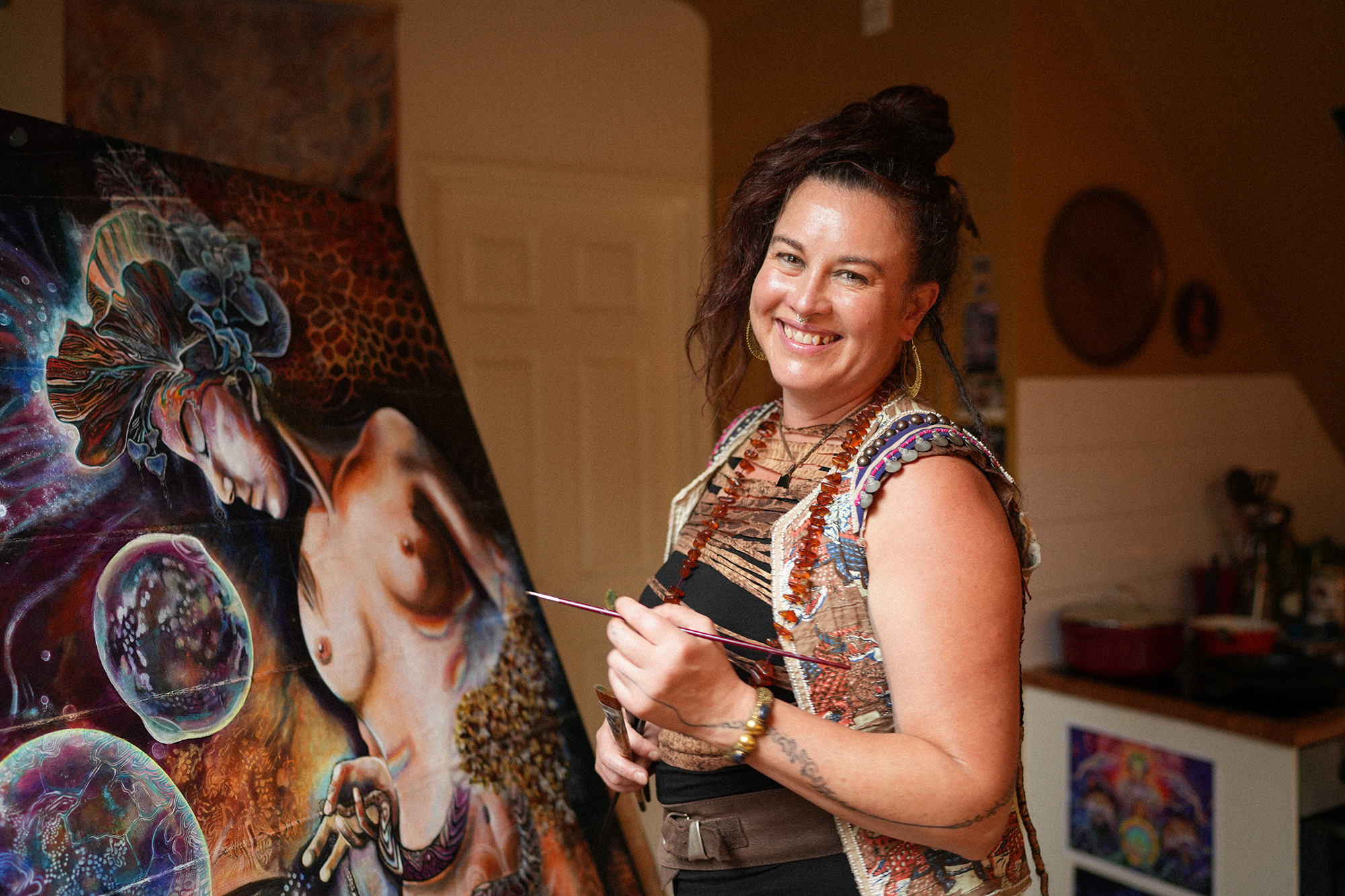 Photo: Barbora Zelenkova In her studio, nestled among her many inspirations, Melissa breathes new life into her large artwork, 'DREAM OF ARTEMIS RISING.' This piece, with its storied past, began on a reclaimed canvas, which she meticulously overlaid with oils, egg tempera, and pure pigment glazes. Remarkably, during one of her art shows, this painting was lost and later found rolled up beside a highway. Unfazed by its misadventures, Melissa embraces the piece's tumultuous journey with optimism. She believes that the artwork now carries an even richer history and a deeper soul, making it all the more captivating.